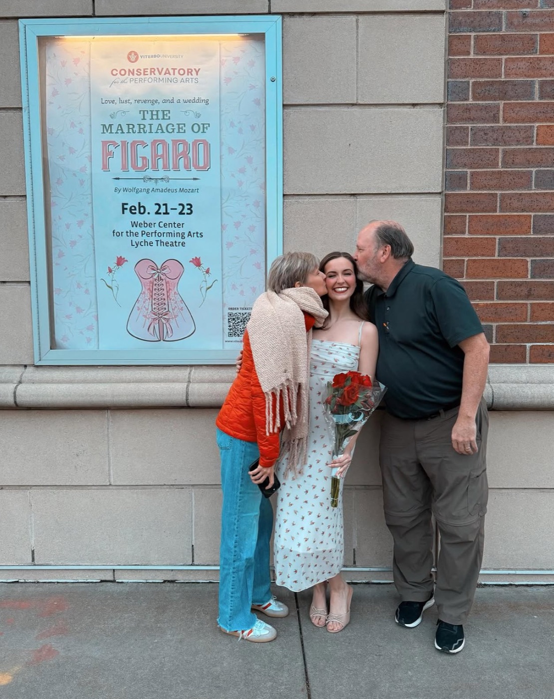 Beth Anderson holding red roses, kissed on both cheeks by her mom and dad beside the poster for The Marriage of Figaro at Viterbo University