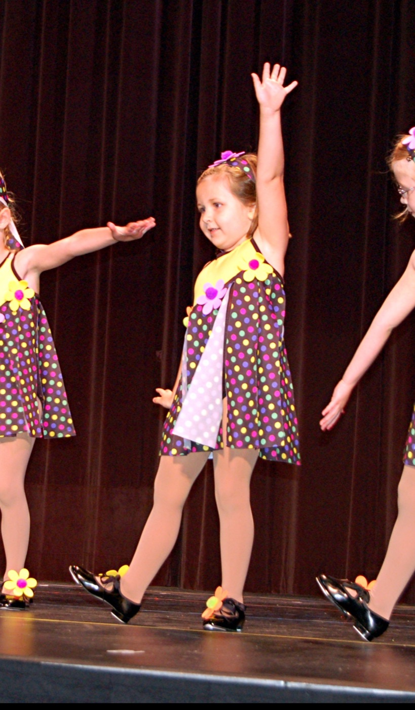 A young Beth Anderson on stage in a polka-dot costume and tap shoes, one arm raised, dancing in a childhood recital