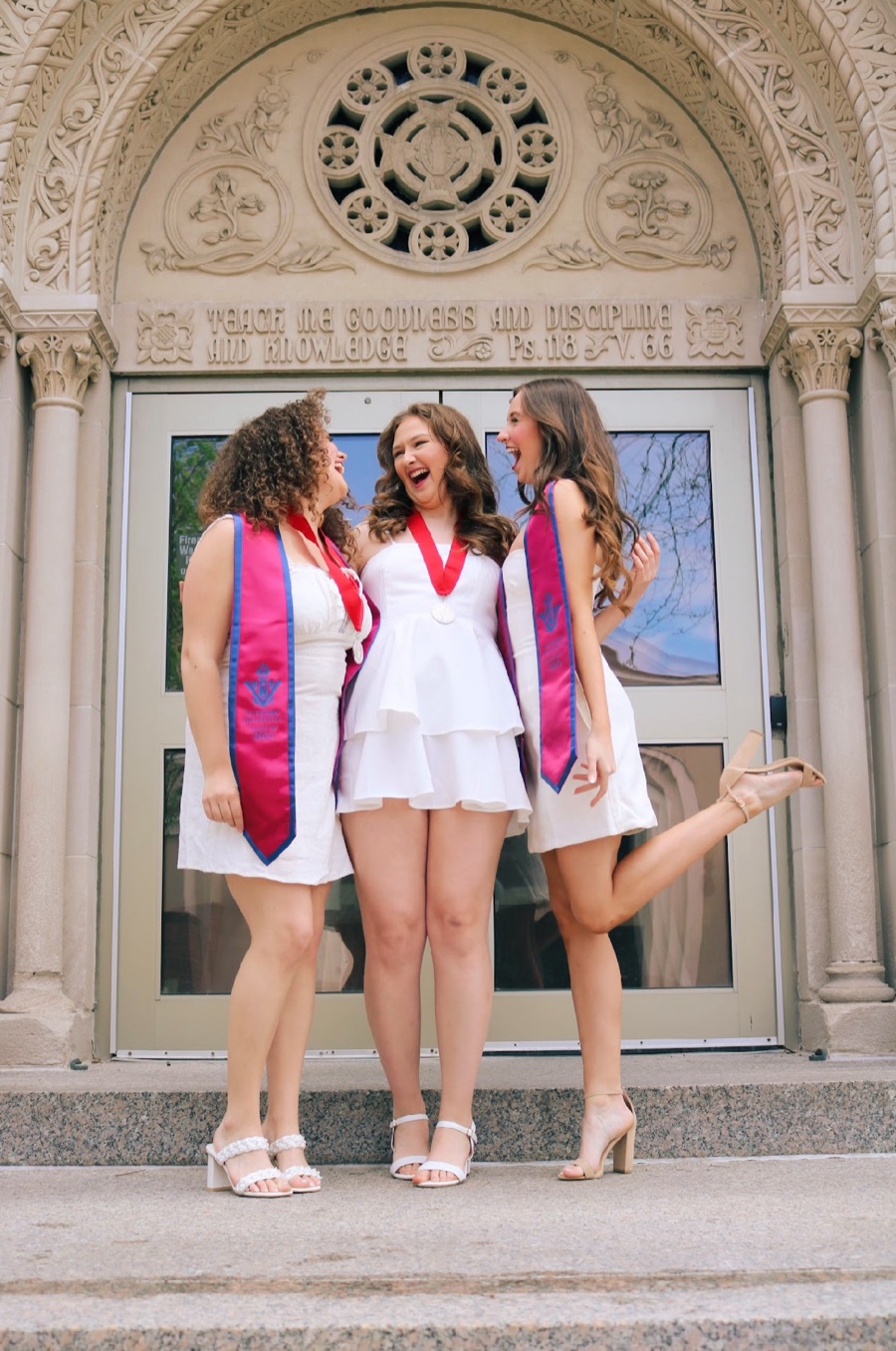 Beth Anderson laughing with two friends on graduation day, all three in white dresses and graduation sashes on the chapel steps