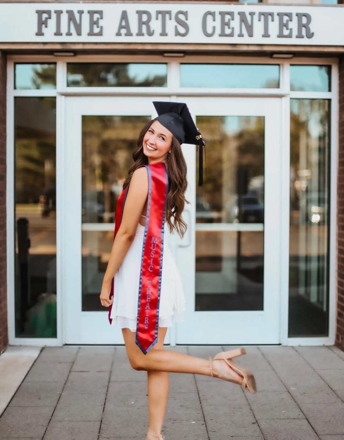 Beth Anderson in cap and gown with a Music Theatre stole, kicking one heel up outside the Fine Arts Center at Viterbo University