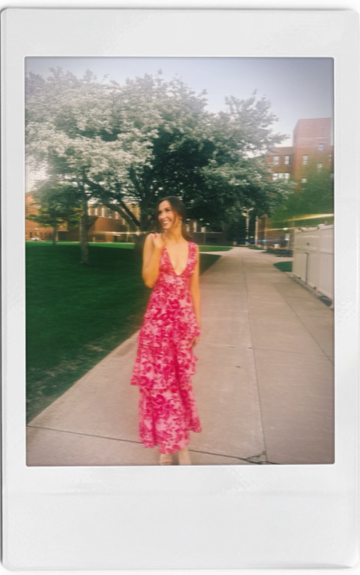 A polaroid of Beth Anderson in a pink and red floral dress, smiling on a campus path under blossoming trees
