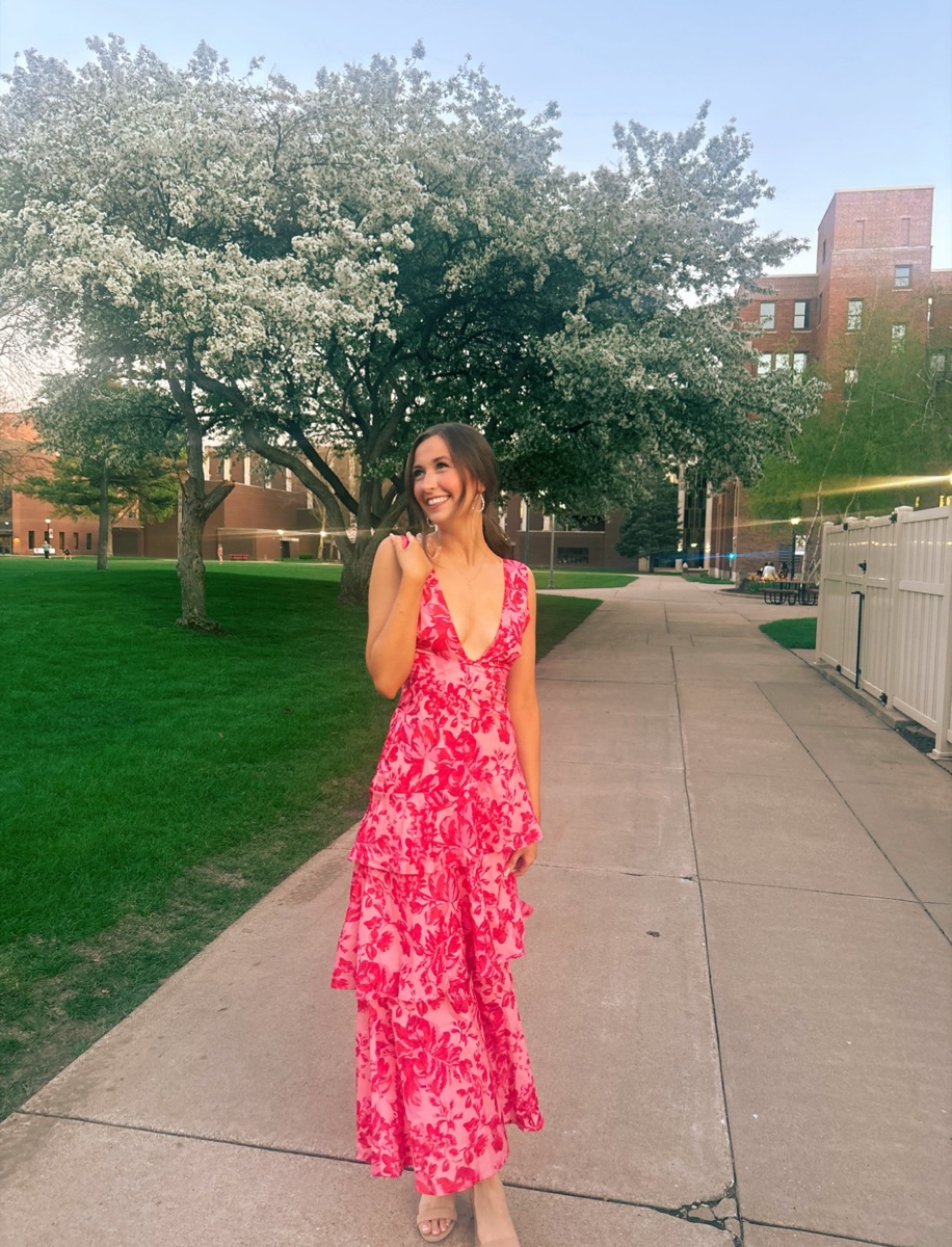 Beth Anderson in a pink and red floral dress, smiling under a blossoming tree on campus at golden hour